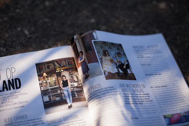 Emilly Prado, a medium skinned woman with black hair is outdoors near a street, reading from a paper while holding a microphone. She wears a hot pink tank top. A small audience, PA speakers, and yellow and red table cloth are in the foreground.