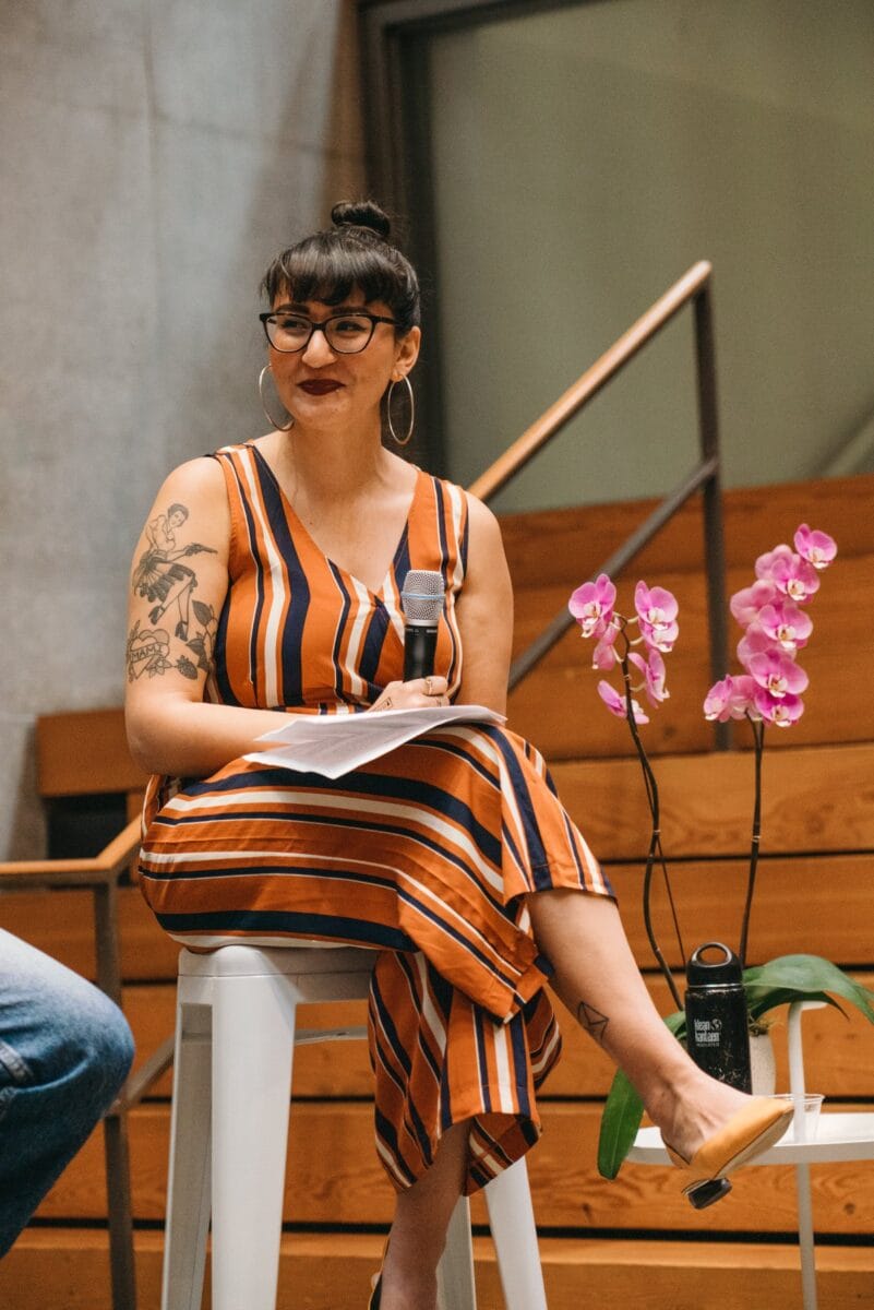Emilly Prado, a medium skinned Latina woman, sits on a stool holding a microphone with a stack of papers in her lap. She sits cross-legged and wears a striped jumpsuit.