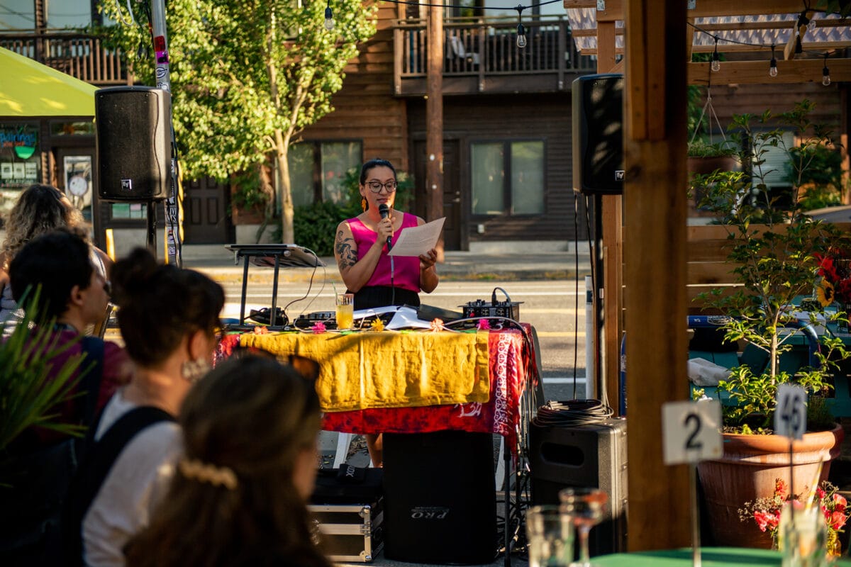 Emilly Prado, a medium skinned woman with black hair is outdoors near a street, reading from a paper while holding a microphone. She wears a hot pink tank top. A small audience, PA speakers, and yellow and red table cloth are in the foreground.