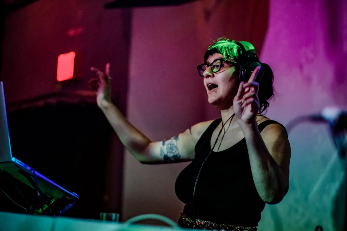 DJ Mami Miami, a female DJ, is singing while in front of a laptop, pointing both hands in the air. She wears headphones, glasses, and a black top with a shiny gold belt made up of numerous cheetahs. Event lights wash her hair in a green tint and in the background is vibrant purples, blue, and red.
