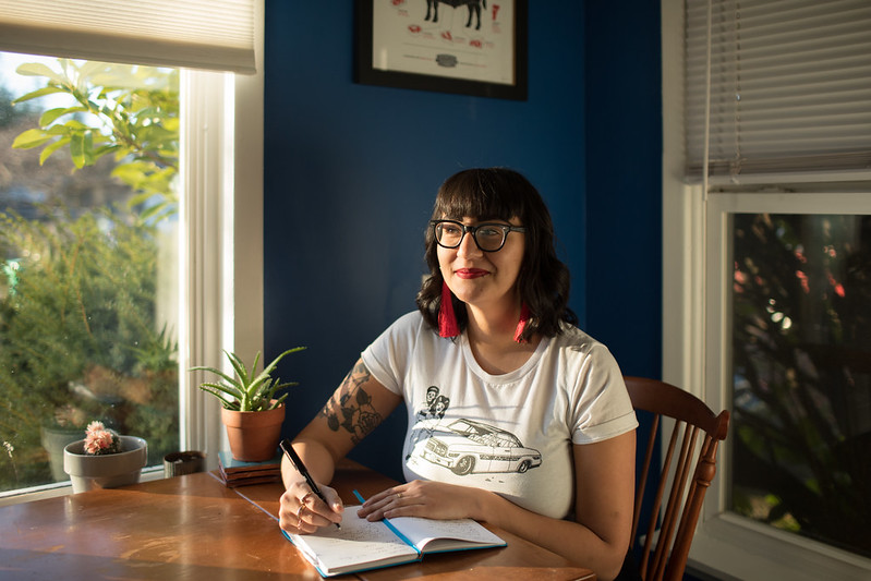 Emilly Prado, a Chicana woman, smiles at a wooden kitchen table writing in a notebook and in front of a window. She wears glasses, her hair curled and shoulder length, and is smiling with closed lips and bright red lipstick. Potted plants, outdoor shrubs, and a bright blue painted wall are in the distance.