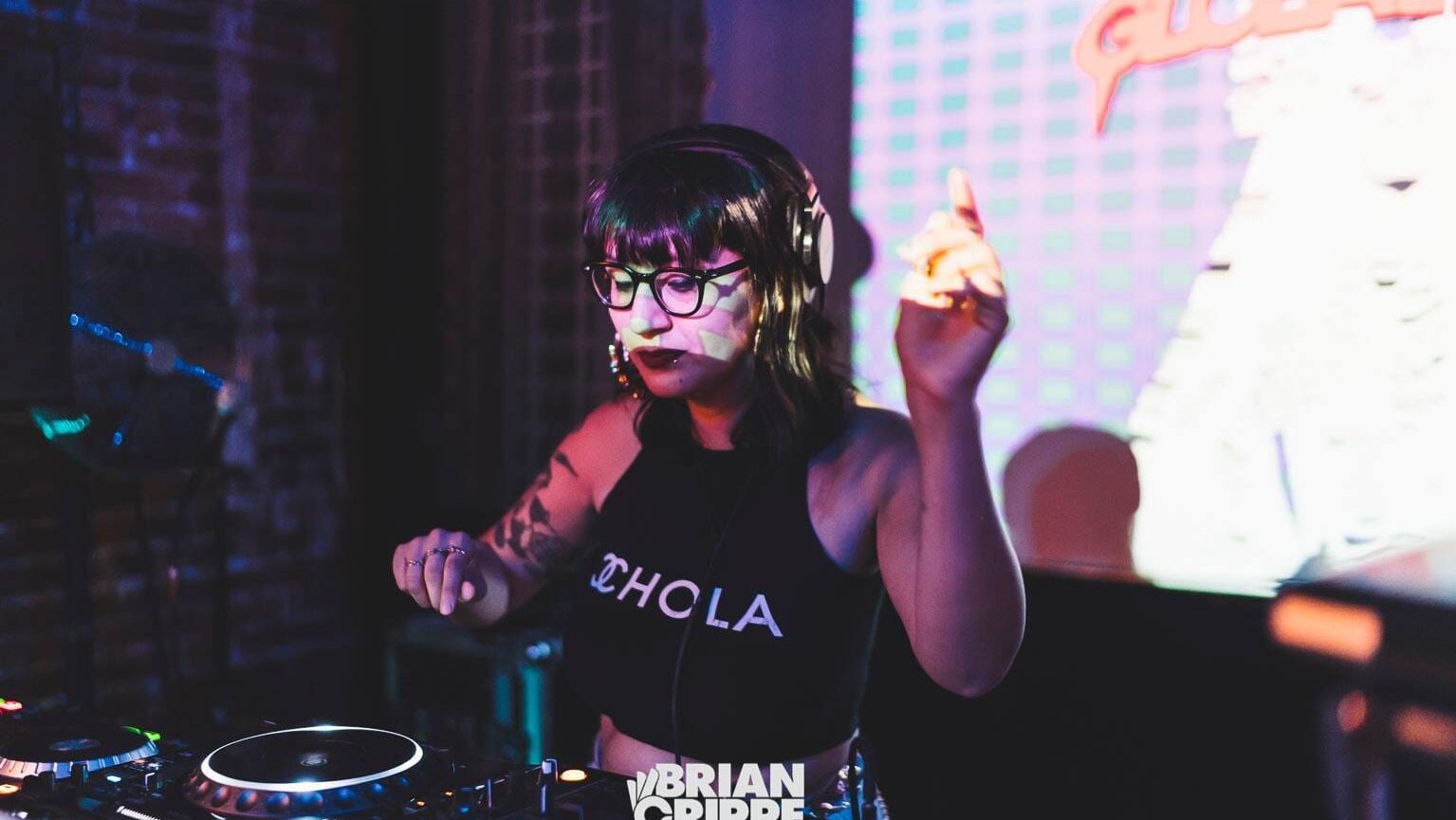 DJ Mami Miami, a medium skinned Latina woman with gold hoop earrings, brown lipstick, and shoulder length hair performing on a DJ controller. Her shirt says "Chola".