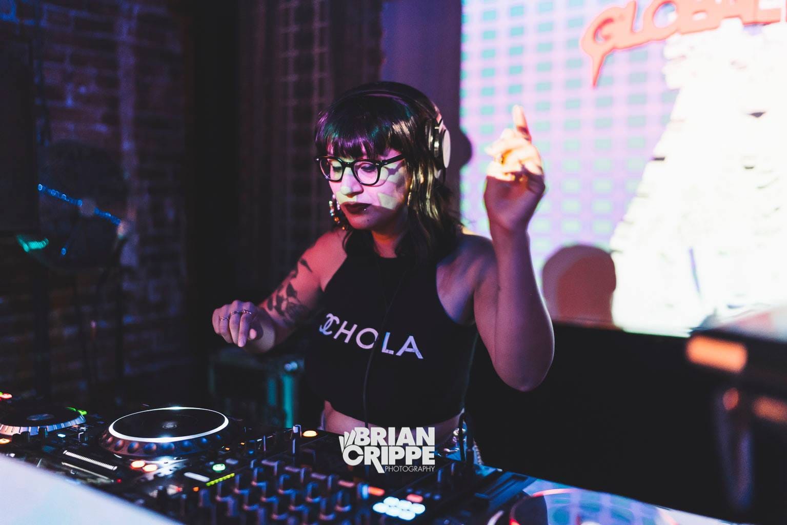 DJ Mami Miami, a medium skinned Latina woman with gold hoop earrings, brown lipstick, and shoulder length hair performing on a DJ controller. Her shirt says "Chola".