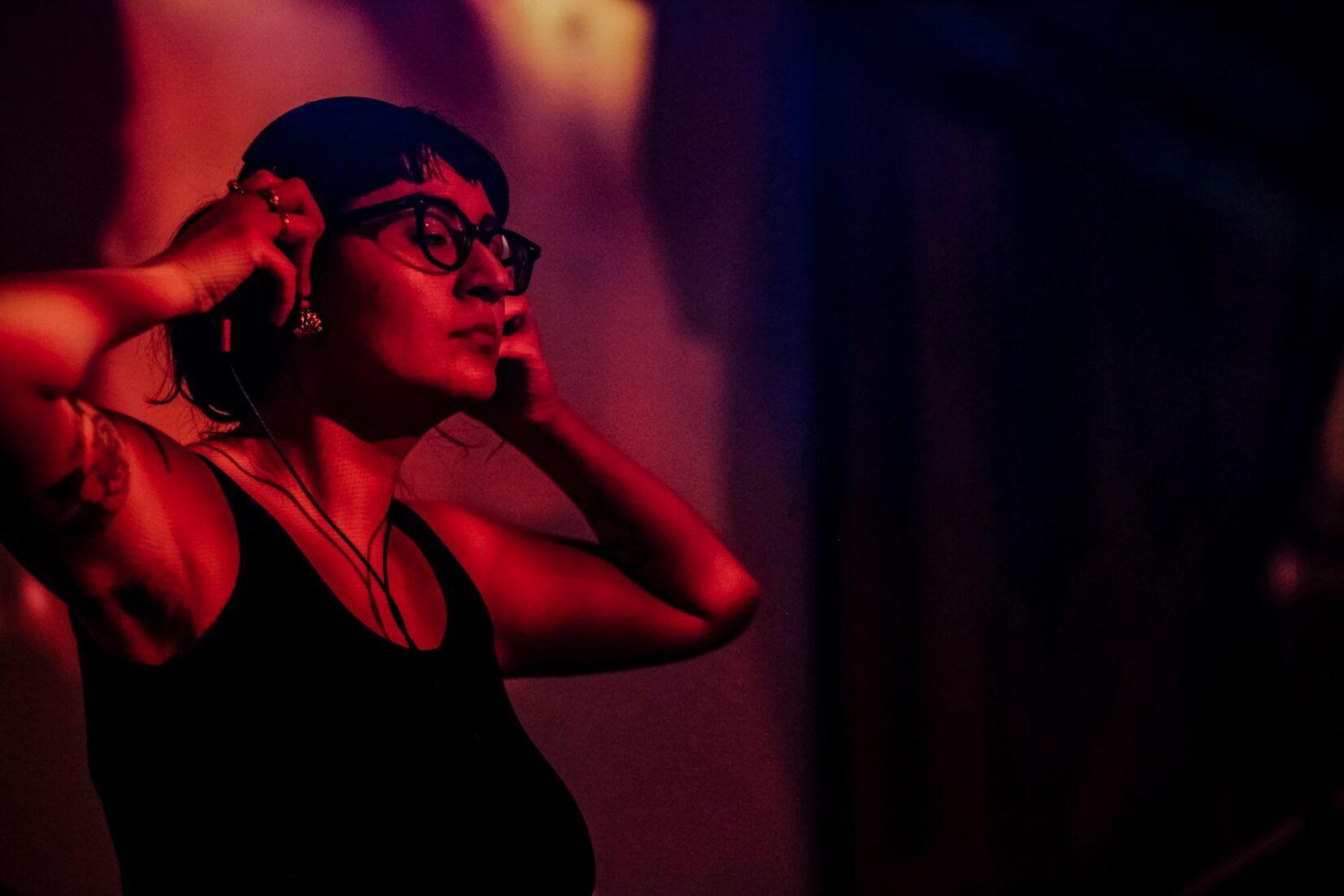 an image of DJ Mami Miami a woman with a pony tail, glasses, and headphones performing with eyes closed, holding her headphones. There is a warm red light wash over the image.