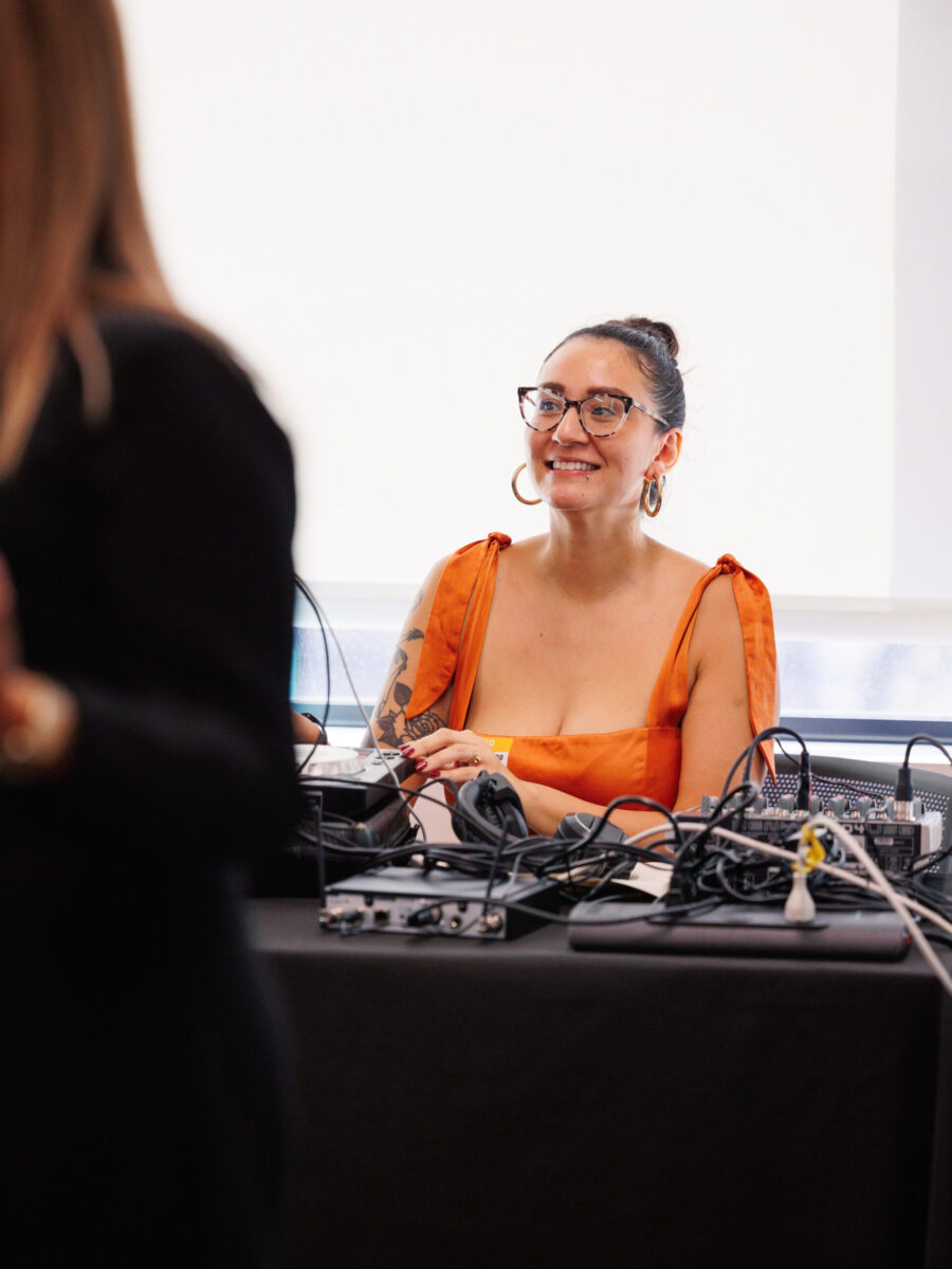 Emilly Prado, a Latina woman sitting and wearing glasses. She is smiling with a sleek bun, orange dress with large ties on the shoulders, and a DJ controller in front of her.