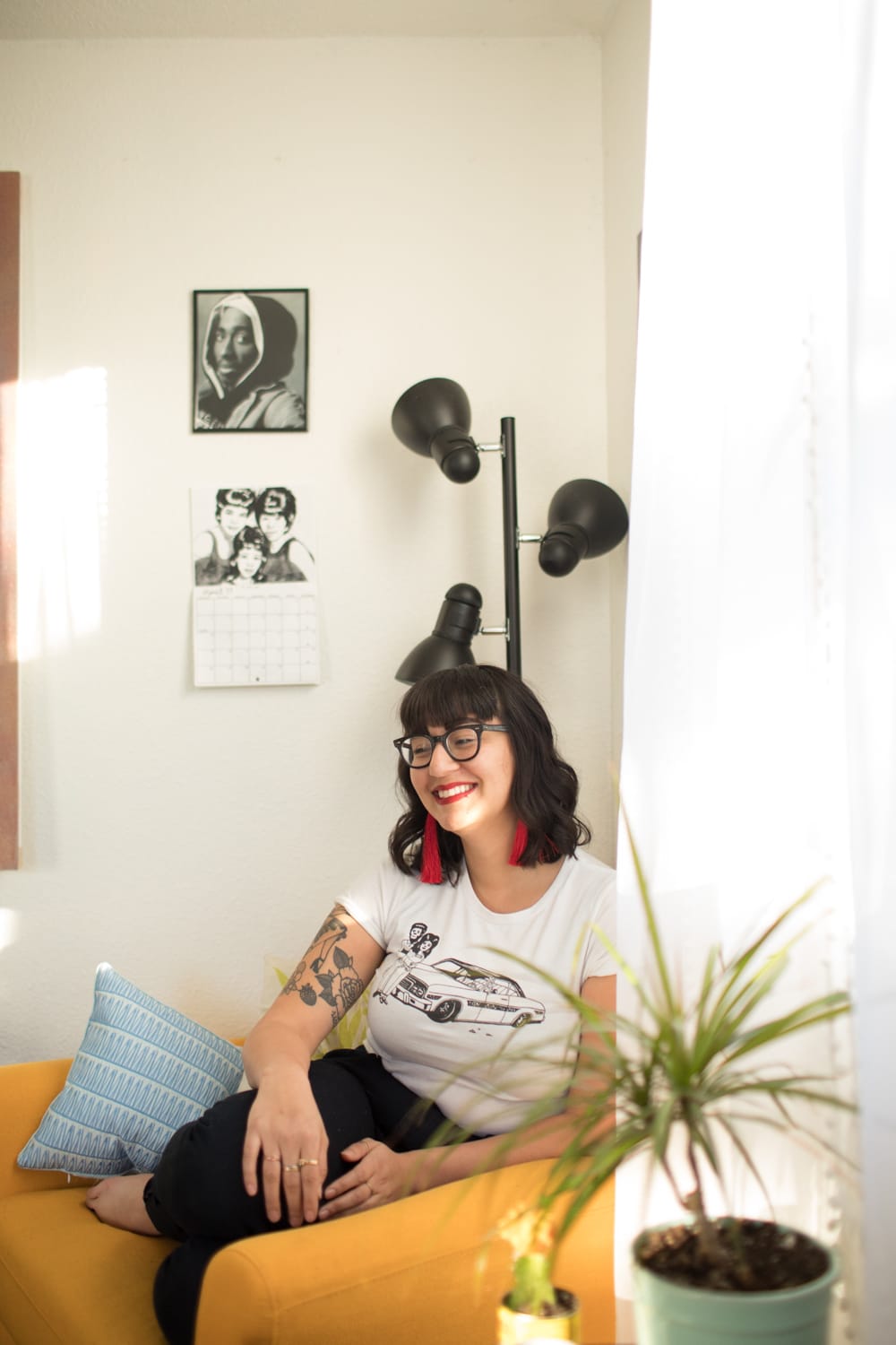 Photo of Emilly Prado, a medium skinned Latina woman, smiling with a downward gaze. She has red lipstick on and sits on a yellow arm chair in a room with bright sunlight, plants, and musician art work.
