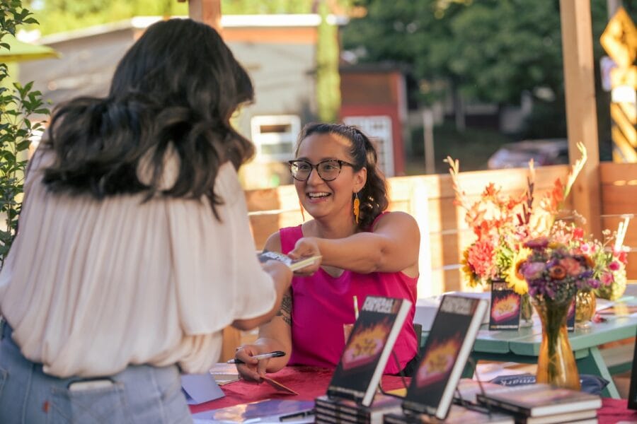 Author Emily Prado, wearing a hot pink shirt and flame earrings, hands over a copy of her book to a reader.