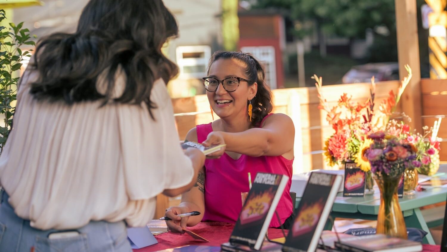 Author Emily Prado, wearing a hot pink shirt and flame earrings, hands over a copy of her book to a reader.