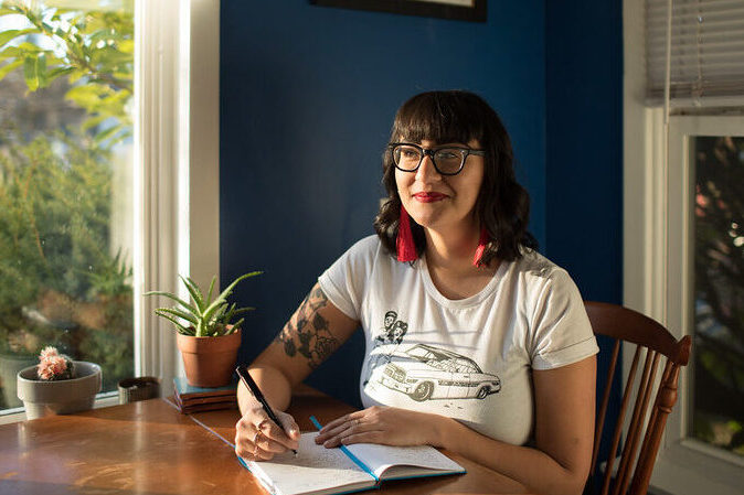 Emilly Prado, a Chicana woman, smiles at a wooden kitchen table writing in a notebook and in front of a window. She wears glasses, her hair curled and shoulder length, and is smiling with closed lips and bright red lipstick. Potted plants, outdoor shrubs, and a bright blue painted wall are in the distance.